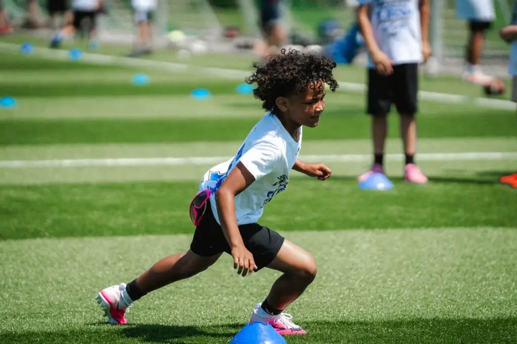 Kind absolviert eine Lauf- und Richtungswechselübung zwischen Markierungshütchen auf einem Kunstrasenplatz während eines American-Football-Camps.