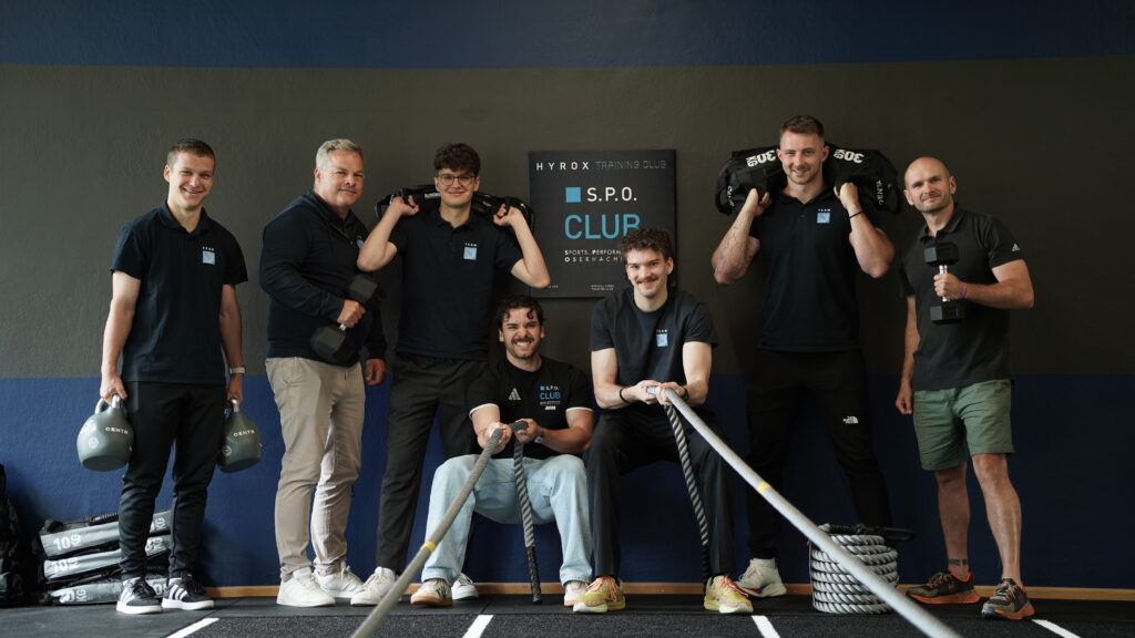 Team des S.P.O Clubs in Oberhaching posiert mit Fitness-Equipment wie Kettlebells, Battle Ropes, Kurzhanteln und Sandbags vor dem Club-Logo.&ldquo;