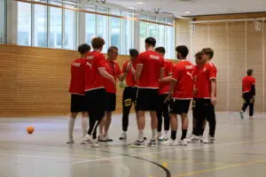 Die U20-Handball-Nationalmannschaft bespricht mit dem Trainer eine Trainingseinheit in einer Sporthalle der Sportschule Oberhaching. Die Spieler tragen rote Deutschland-Trikots und stehen im Kreis auf dem Hallenboden.