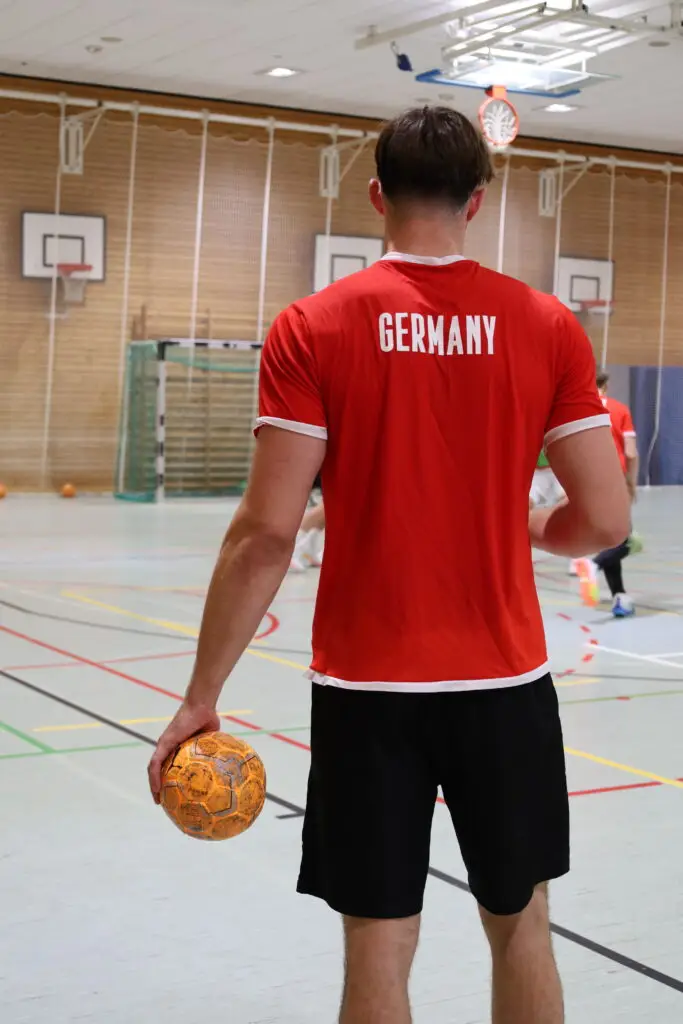 Ein Spieler der U20-Handball-Nationalmannschaft steht mit einem Handball in der Hand in einer Sporthalle der Sportschule Oberhaching. Er tr&auml;gt ein rotes Deutschland-Trikot mit der Aufschrift &bdquo;GERMANY&ldquo; auf dem R&uuml;cken, w&auml;hrend im Hintergrund weitere Spieler trainieren.