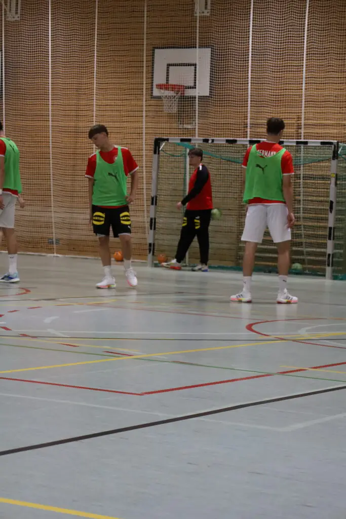 Spieler der U20-Handball-Nationalmannschaft absolvieren eine Trainingseinheit in einer Sporthalle der Sportschule Oberhaching. Einige Athleten tragen gr&uuml;ne Leibchen, w&auml;hrend im Hintergrund ein Torwart vor dem Handballtor steht.