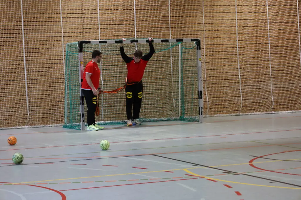 Ein Trainer unterst&uuml;tzt einen Spieler der U20-Handball-Nationalmannschaft bei einer &Uuml;bung mit Widerstandsband am Handballtor in einer Sporthalle der Sportschule Oberhaching. Mehrere Handb&auml;lle liegen auf dem Hallenboden bereit.