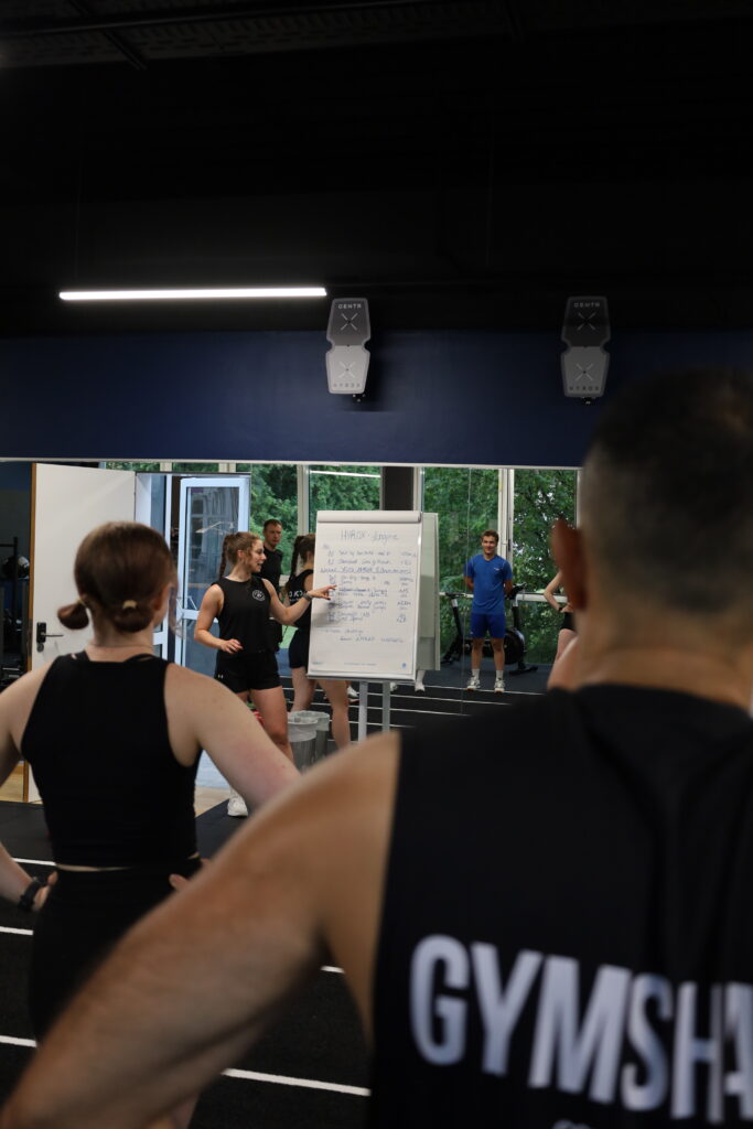 Trainerin erkl&auml;rt den Ablauf eines HYROX-Workouts im S.P.O. Club der Sportschule Oberhaching vor einer Gruppe von Teilnehmenden.