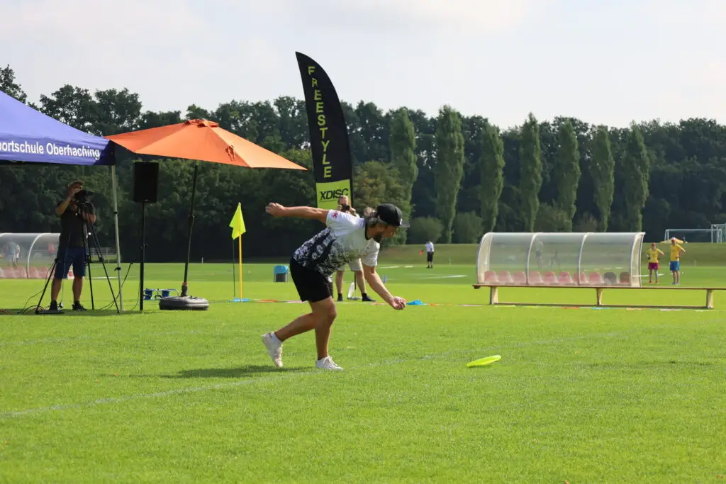 Frisbee-Spieler beim Wurf auf dem Sportplatz der Sportschule Oberhaching während des Frisbee Festivals, mit Freestyle-Banner und Zuschauenden im Hintergrund.