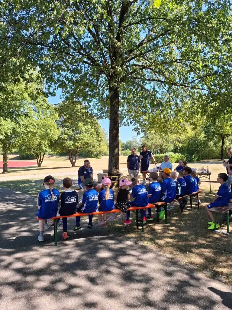 Kinder in blauen BFV-Trikots sitzen bei einem Fußball-Aktionstag der BFV-Sozialstiftung und des Ambulanten Kinderhospizes auf Bierbänken im Schatten, während ein Trainer an der Sportschule Oberhaching mit ihnen spricht.