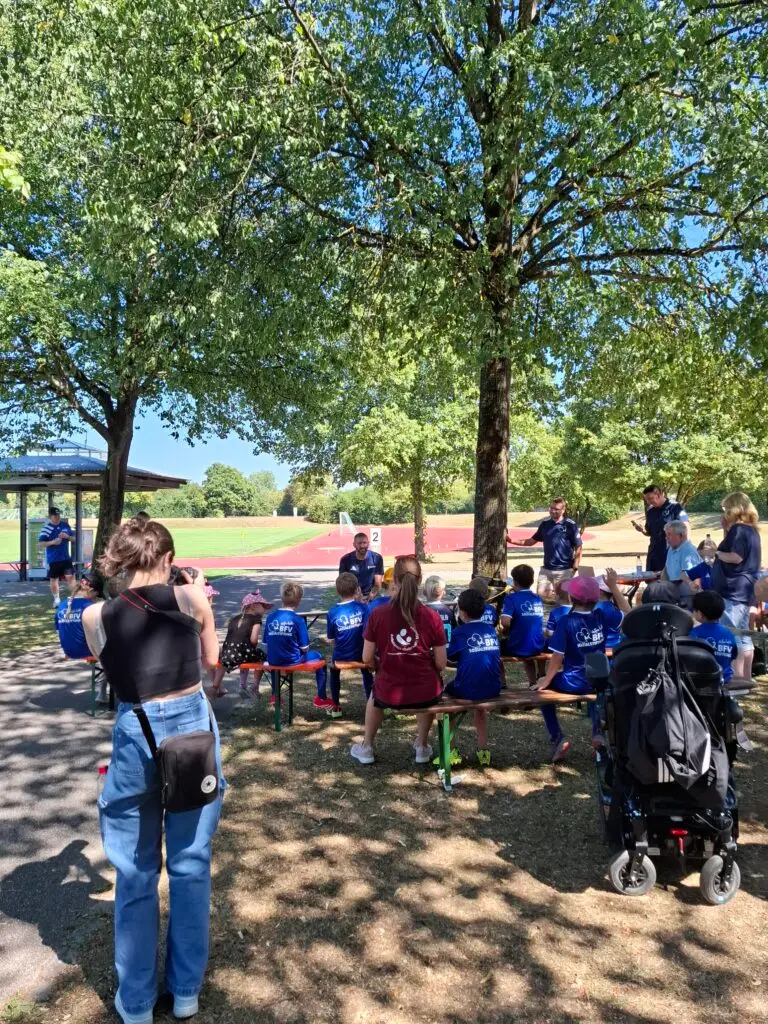 Kinder in blauen BFV-Trikots sitzen bei einem Fußball-Aktionstag der BFV-Sozialstiftung und des Ambulanten Kinderhospizes auf Bierbänken im Schatten, während ein Trainer an der Sportschule Oberhaching mit ihnen spricht.