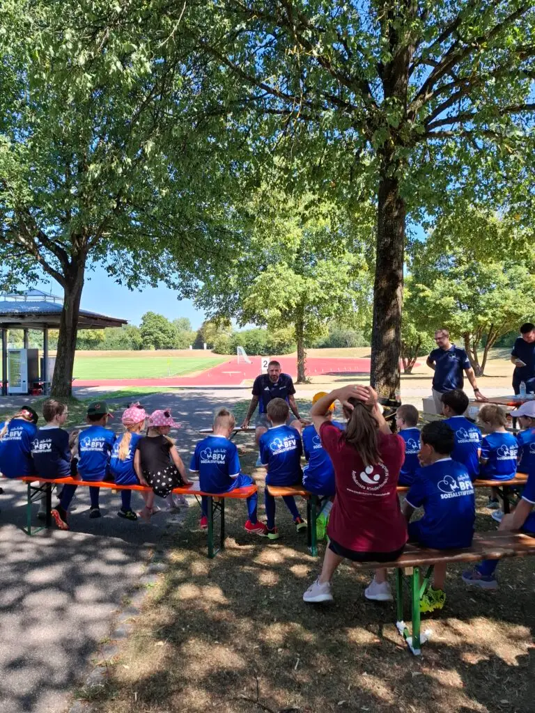 Kinder in blauen BFV-Trikots sitzen bei einem Fußball-Aktionstag der BFV-Sozialstiftung und des Ambulanten Kinderhospizes auf Bierbänken im Schatten, während ein Trainer an der Sportschule Oberhaching mit ihnen spricht.