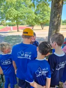 Kinder in blauen BFV-Sozialstiftung-Trikots stehen bei einem Fußball-Aktionstag im Schatten zusammen und sprechen mit einem Betreuer an der Sportschule Oberhaching