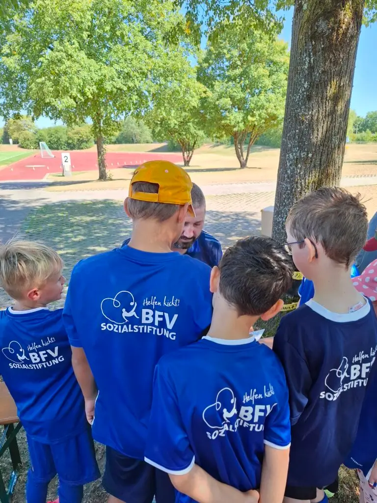 Kinder in blauen BFV-Sozialstiftung-Trikots stehen bei einem Fußball-Aktionstag im Schatten zusammen und sprechen mit einem Betreuer an der Sportschule Oberhaching