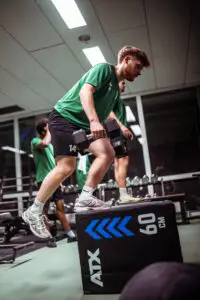 Athlet absolviert im Kraftraum der Sportschule Oberhaching eine Kraft- und Stabilitätsübung mit Kurzhanteln auf einer Plyobox, während weitere Sportler im Hintergrund trainieren.