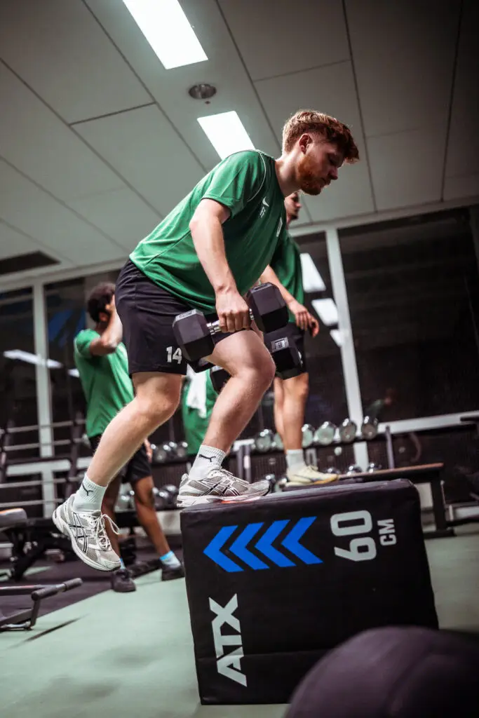 Athlet absolviert im Kraftraum der Sportschule Oberhaching eine Kraft- und Stabilitätsübung mit Kurzhanteln auf einer Plyobox, während weitere Sportler im Hintergrund trainieren.