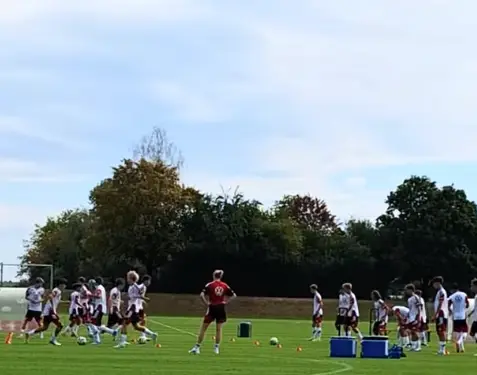 DFB-U16-Nationalmannschaft beim Training auf dem Fußballplatz der Sportschule mit Trainern und Spielern in weißen Trikots. Im Hintergrund sind Bäume und ein bewölkter Himmel zu sehen.